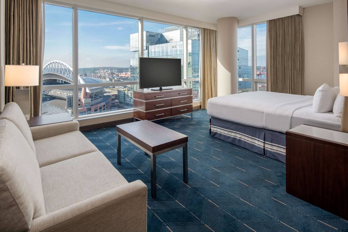 A hotel room with a large bed, a sofa, a TV on a wooden cabinet, and a view of a cityscape through large windows.