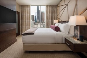 A hotel room with a large bed, a TV, a nightstand with a lamp, and a view of city buildings.