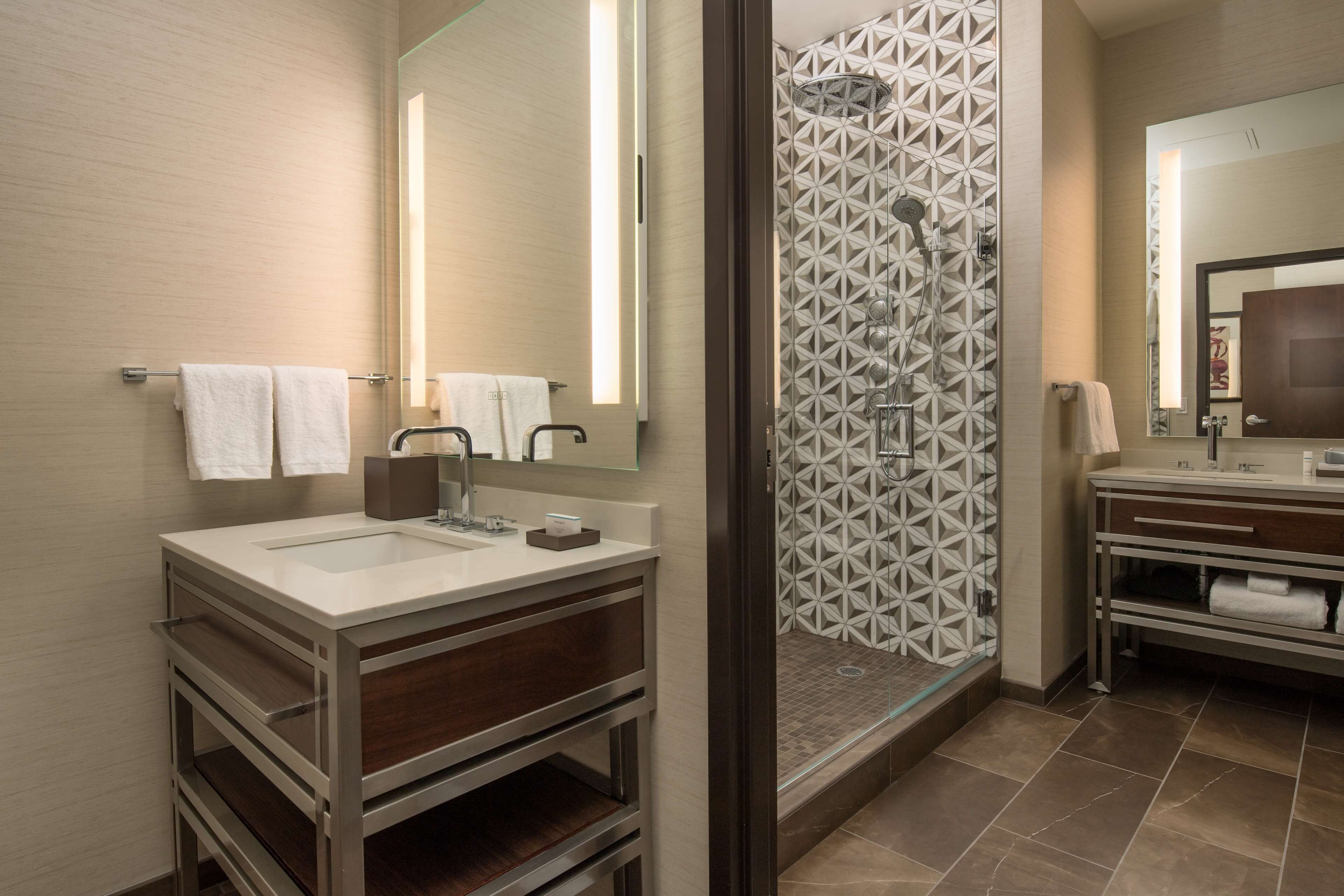Un baño moderno con un espejo grande, dos lavabos, una ducha con azulejos geométricos y un mueble de tocador con espacio de almacenamiento.