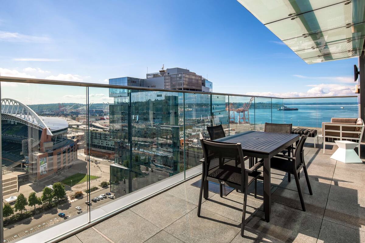 A balcony with a glass railing overlooking a cityscape, including a stadium and water body.