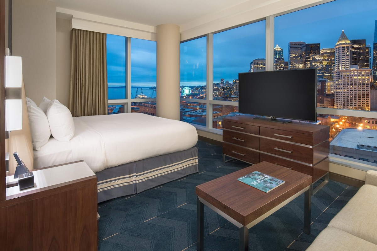 A hotel room with a large bed, a TV, a desk, and a view of the cityscape.