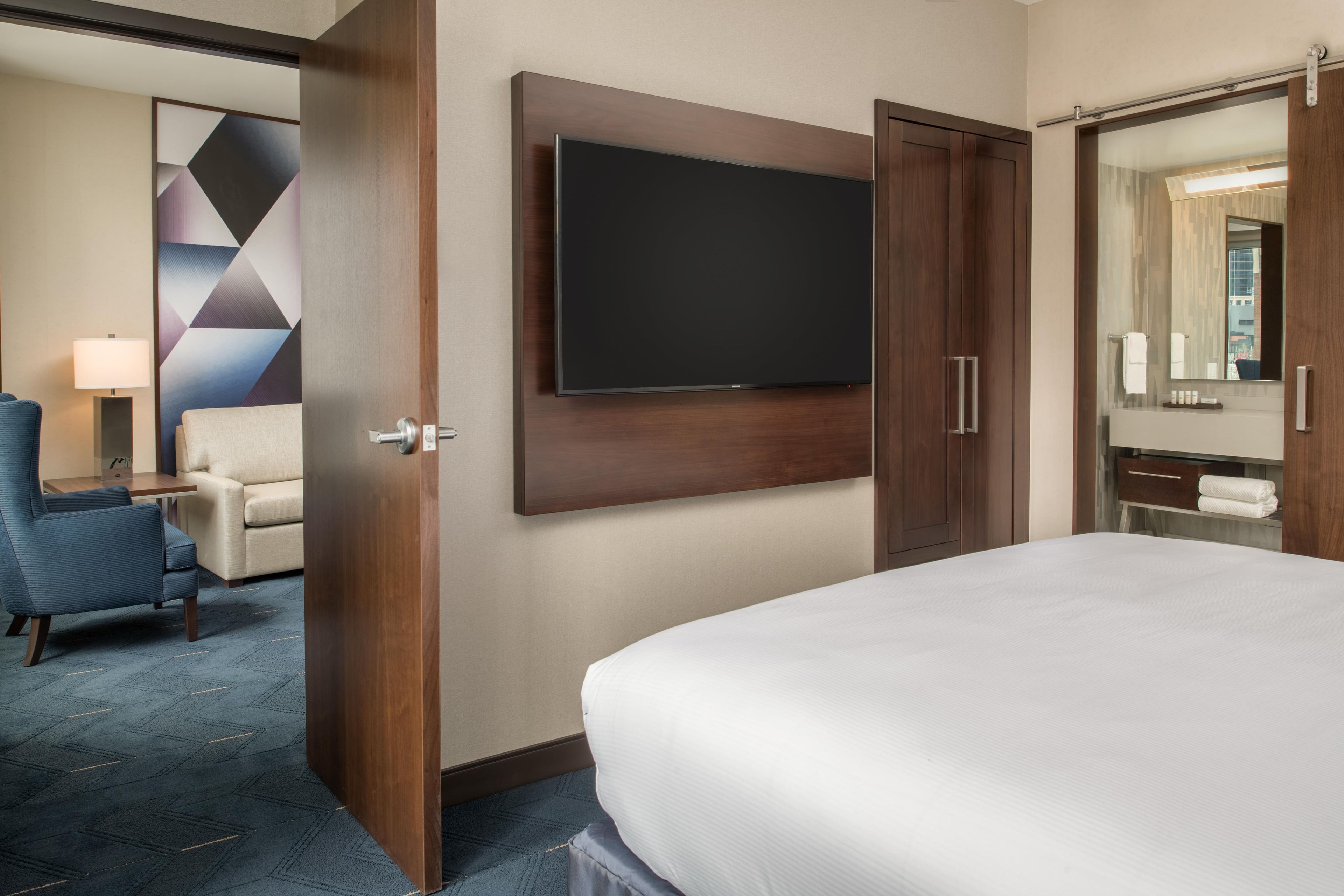 A hotel room with a bed, a TV mounted on the wall, a chair, a desk, and a bathroom with a mirror.