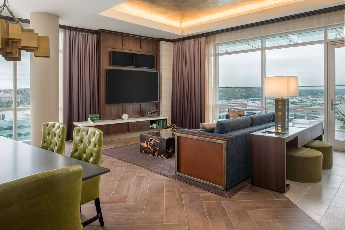 A modern hotel room with a large window, a flat-screen TV, a sofa, a dining table, and a lamp.