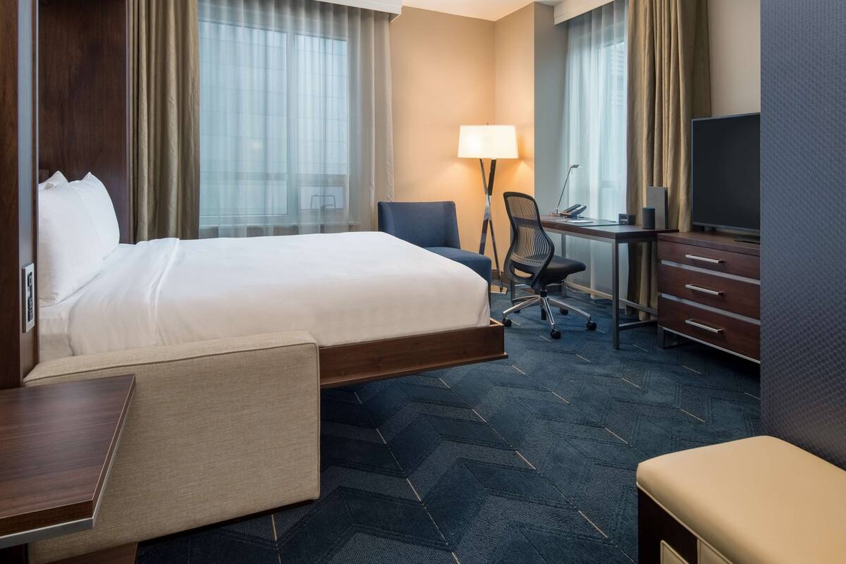 A hotel room with a bed, desk, chair, TV, and a lamp.