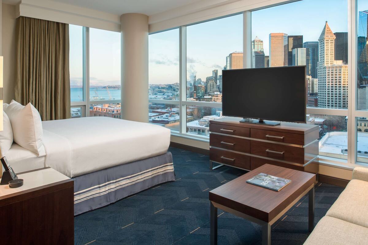 A hotel room with a large bed, a TV, a desk, and a view of a city skyline.
