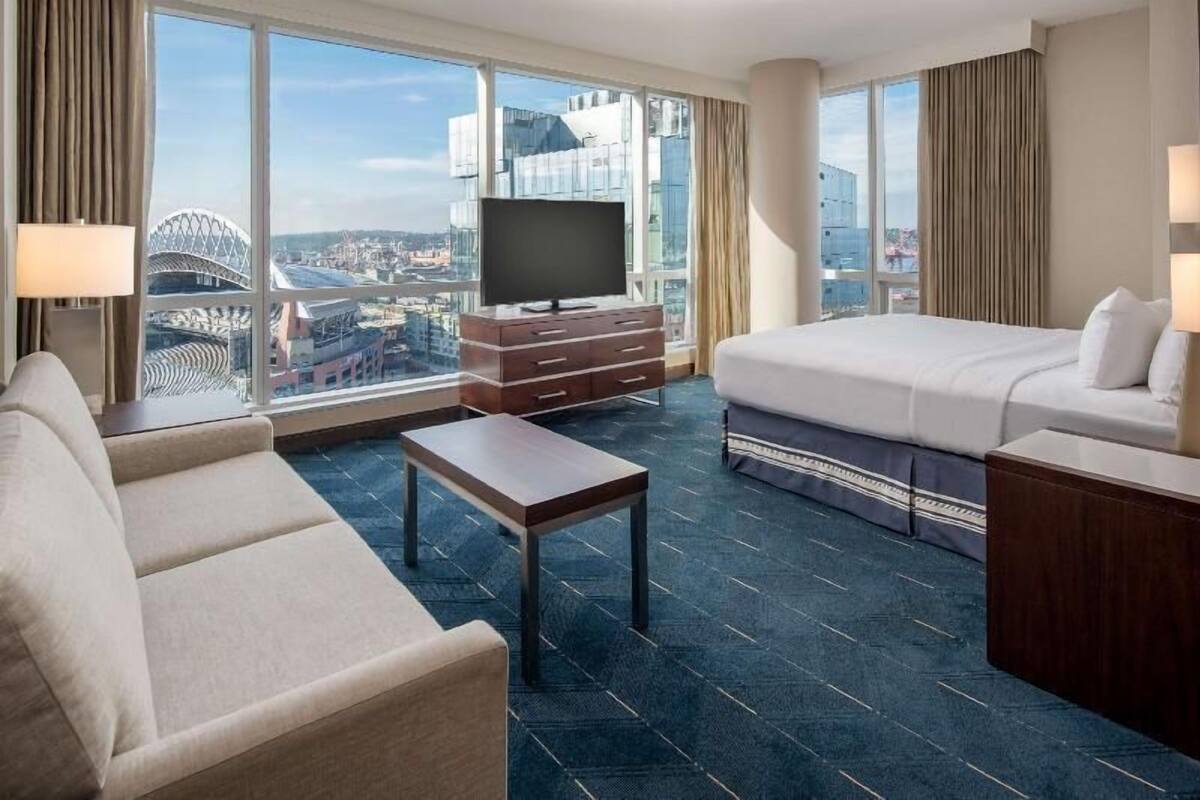 A hotel room with a bed, a sofa, a TV, a desk, and a view of a cityscape.
