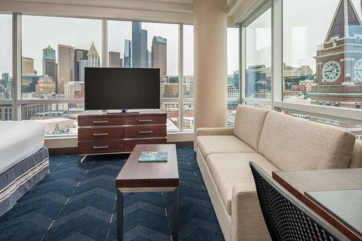 A hotel room with a large window, a flat-screen TV, a sofa, a bed, and a small table.