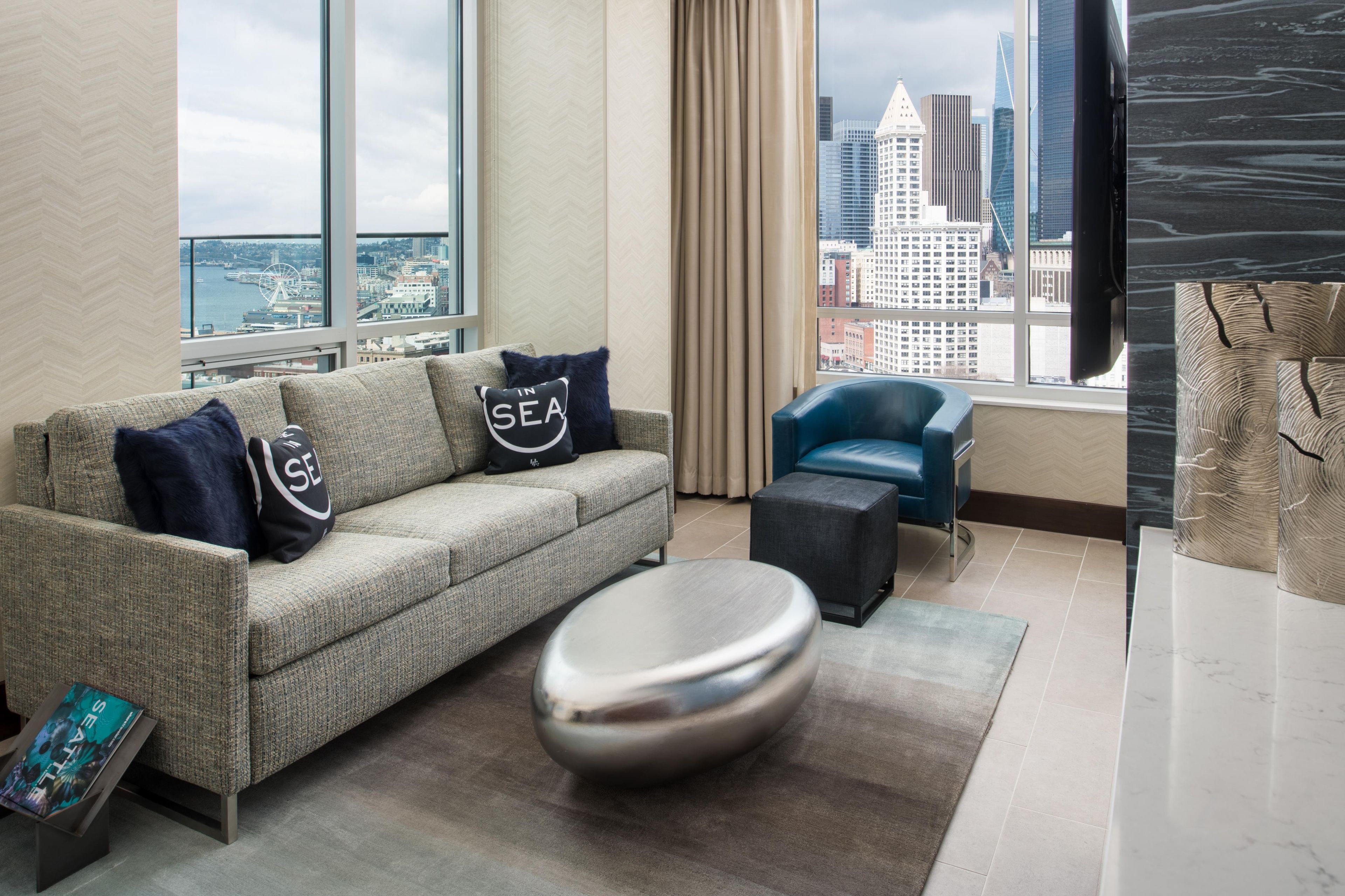 Un salon moderne avec un canapé, un fauteuil bleu, un pouf rond et une vue sur le paysage urbain, visible à travers de grandes fenêtres.