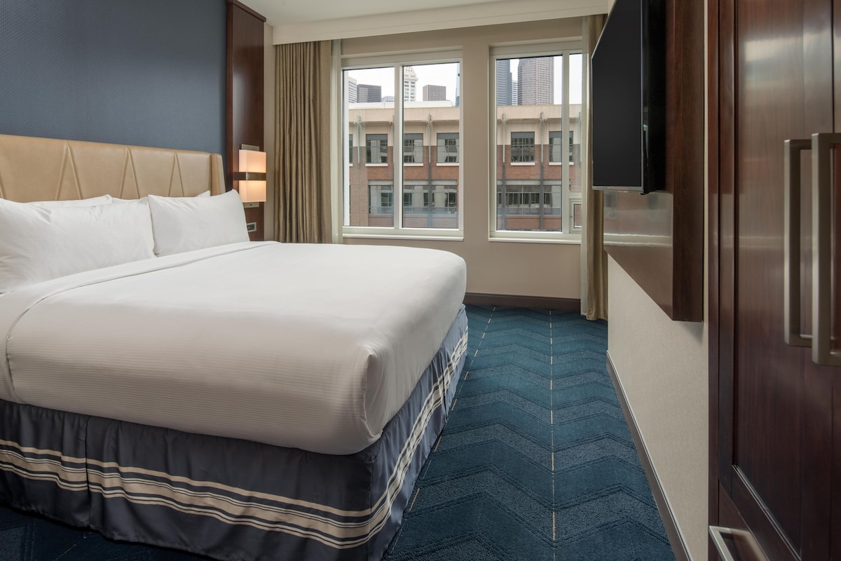 Une chambre d’hôtel avec un grand lit, un téléviseur à écran plat et une vue sur les immeubles par la fenêtre.