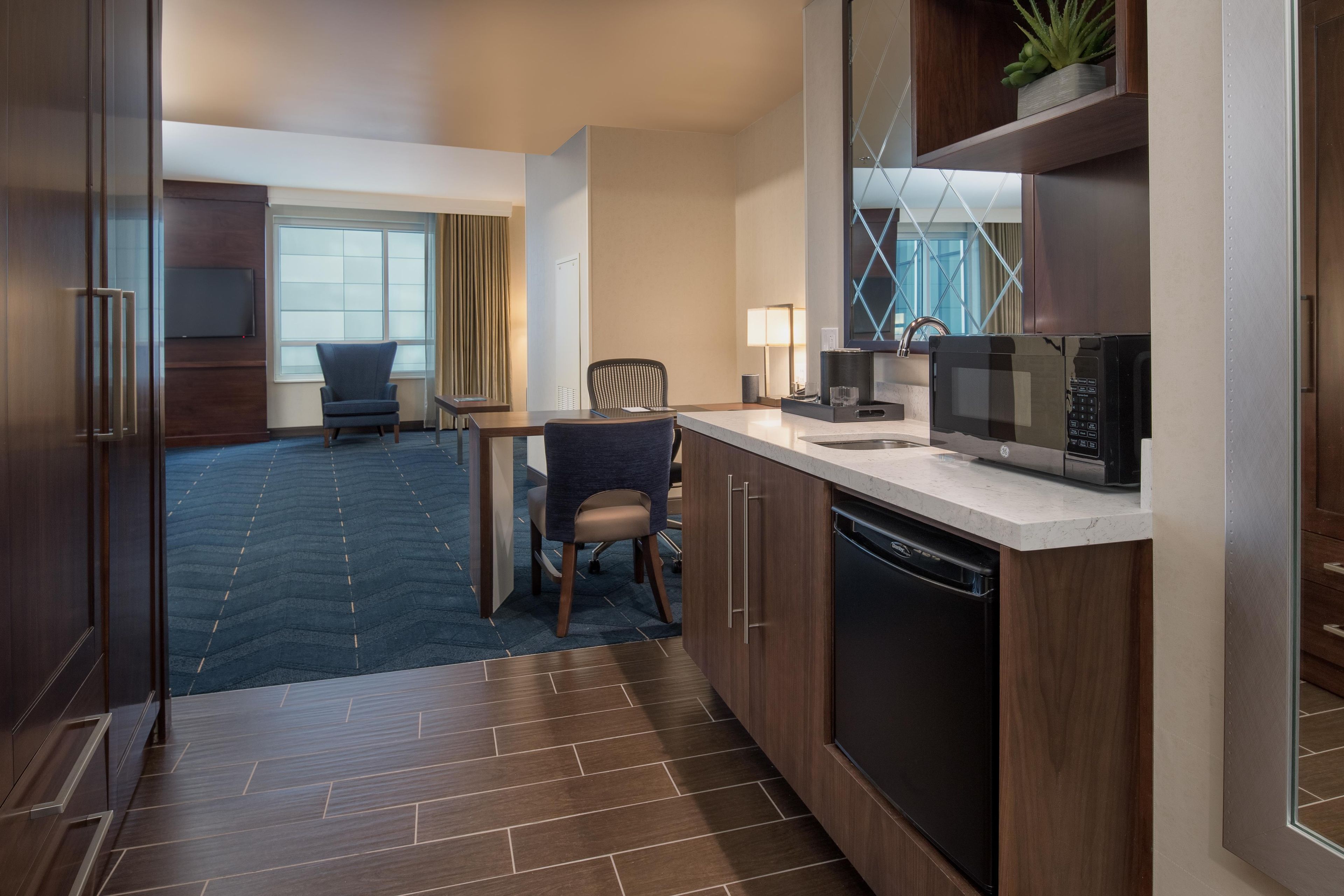 A hotel room with a kitchenette, a dining area with two chairs, a large window with curtains, and a dark wood floor.
