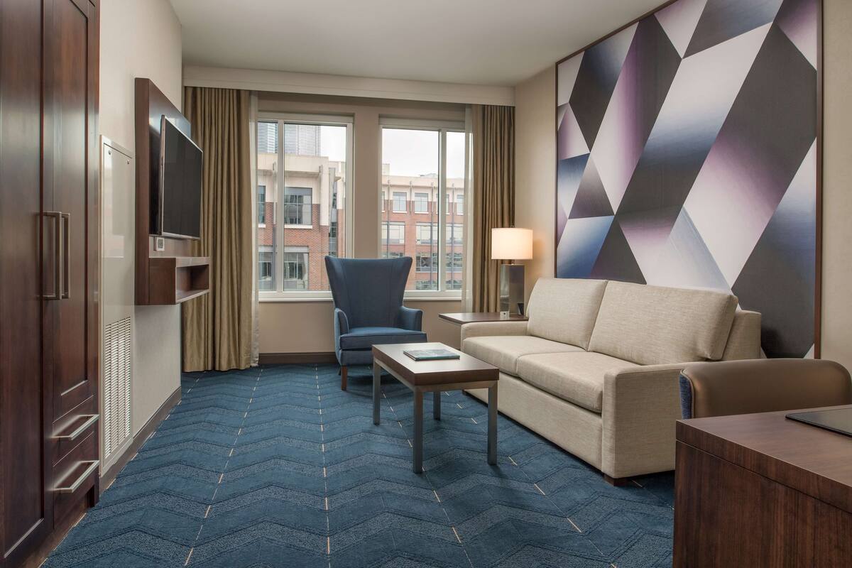 A hotel room with a large window, a sofa, a chair, a small table, and a TV.