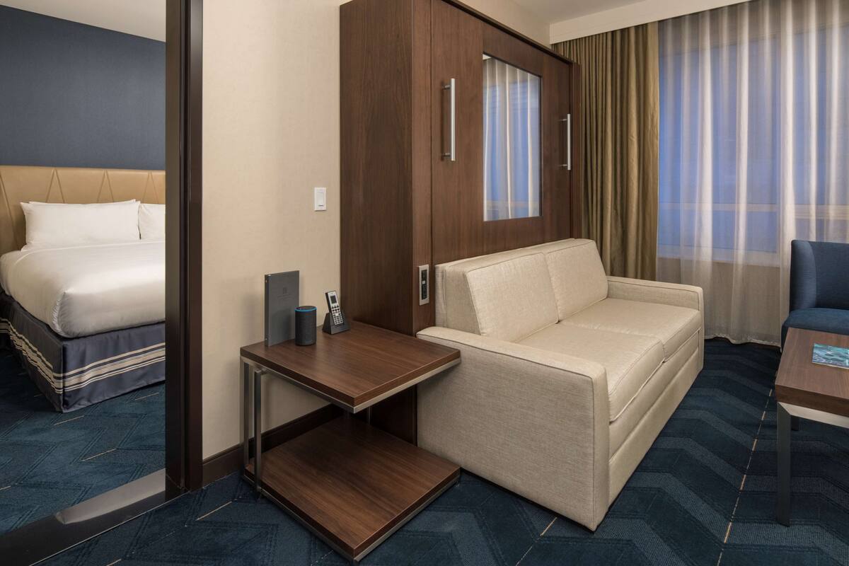 A hotel room with a bed, a sofa, a desk with a phone, and a window with curtains.