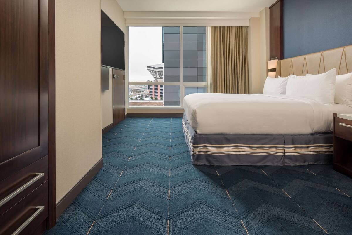 A hotel room with a large bed, a view of a cityscape through a window, a blue carpet, and a wooden wardrobe.