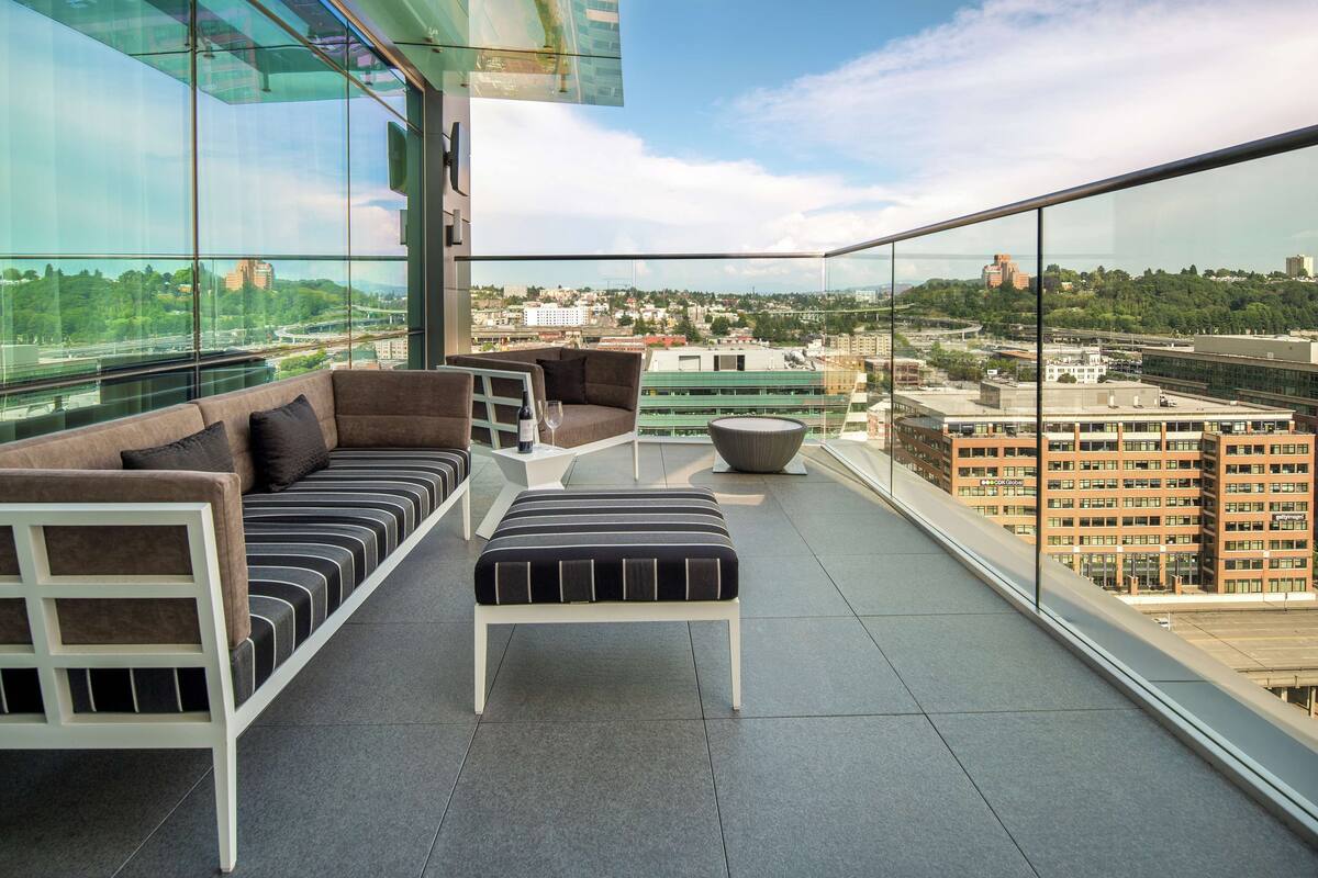 A modern balcony with a sofa, ottoman, and glass railing, offering a city view.
