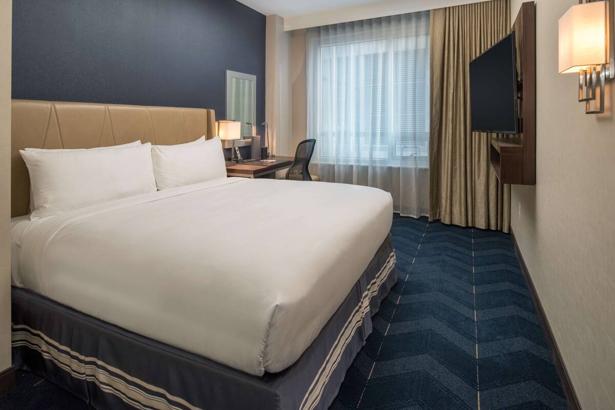 A hotel room with a large bed, a desk with a chair, a TV mounted on the wall, and a window with curtains.
