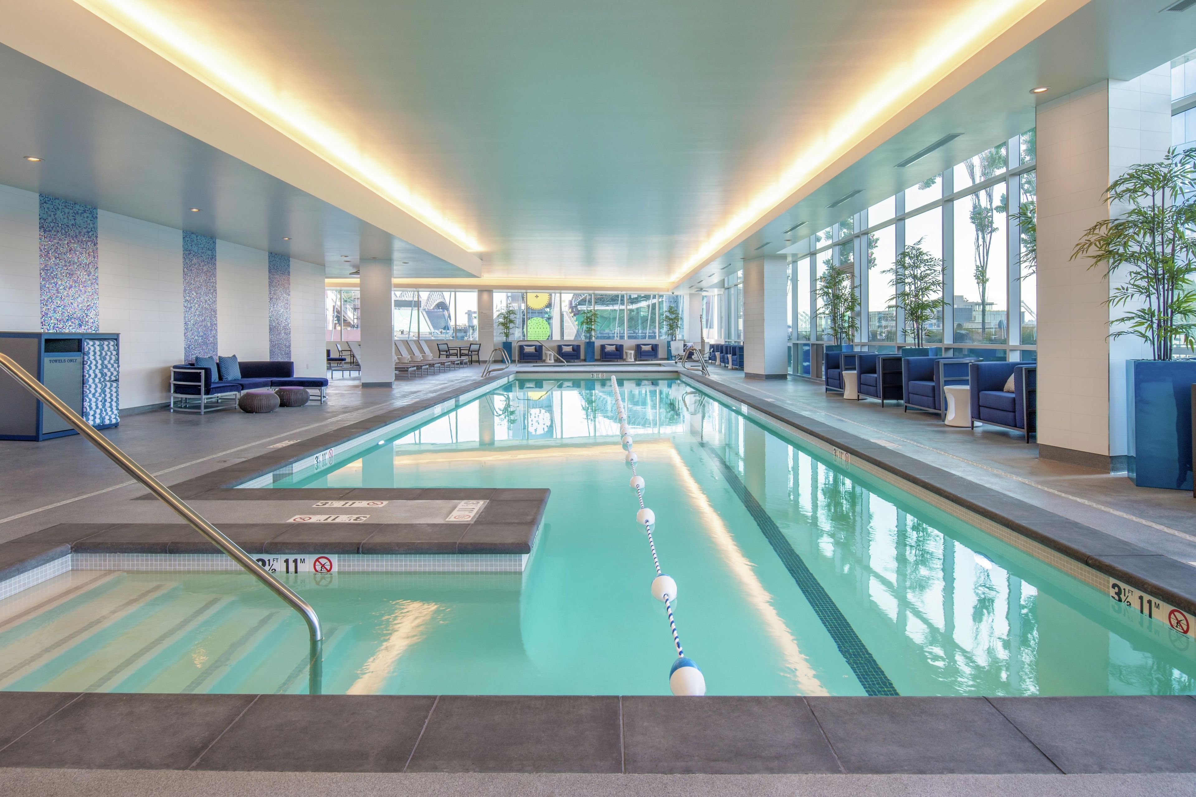 An indoor swimming pool with clear lane markings, a lifeguard stand, and seating areas with a view of the cityscape.