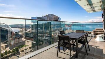 A balcony with a glass railing overlooking a cityscape, a table with chairs, and a view of a stadium and water.