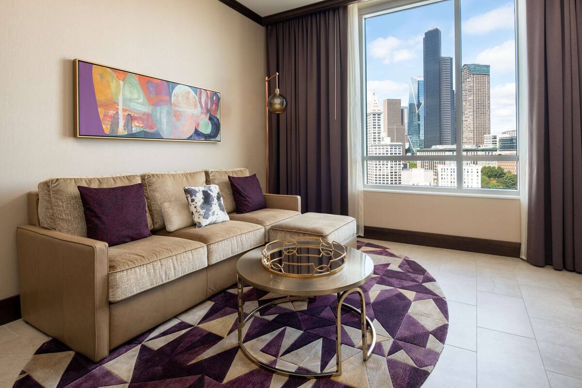 A modern living room with a beige sofa, a patterned rug, and a city view through large windows.