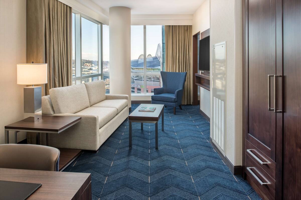 A hotel room with a sofa, a small table, a chair, a TV, and a large window with a view of a Ferris wheel.