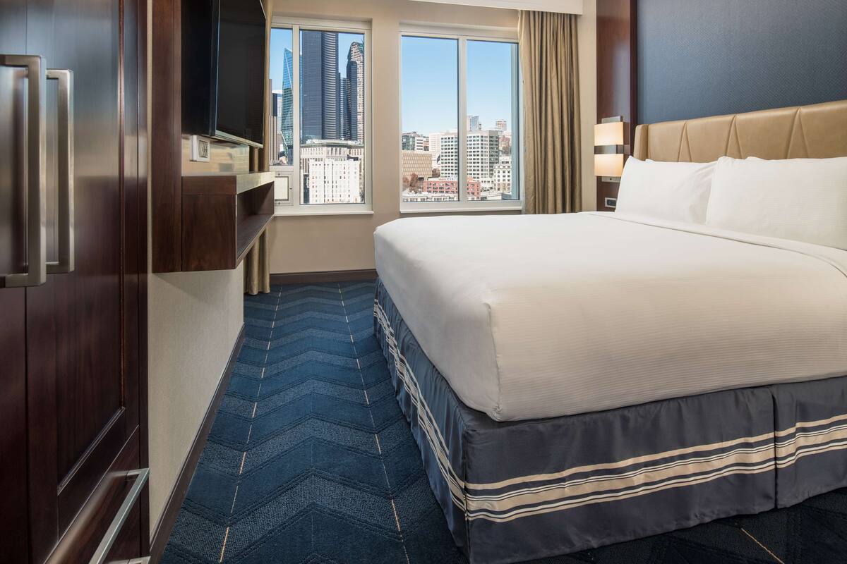 A hotel room with a large bed, a television, a city view, and a blue carpet.
