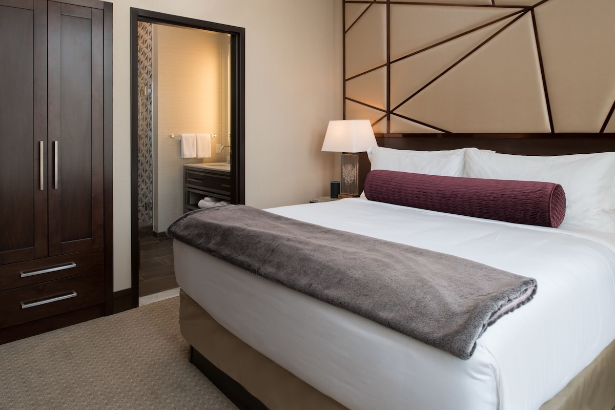 A hotel room with a large bed, a nightstand with a lamp, a wardrobe, and a bathroom visible through an open door.