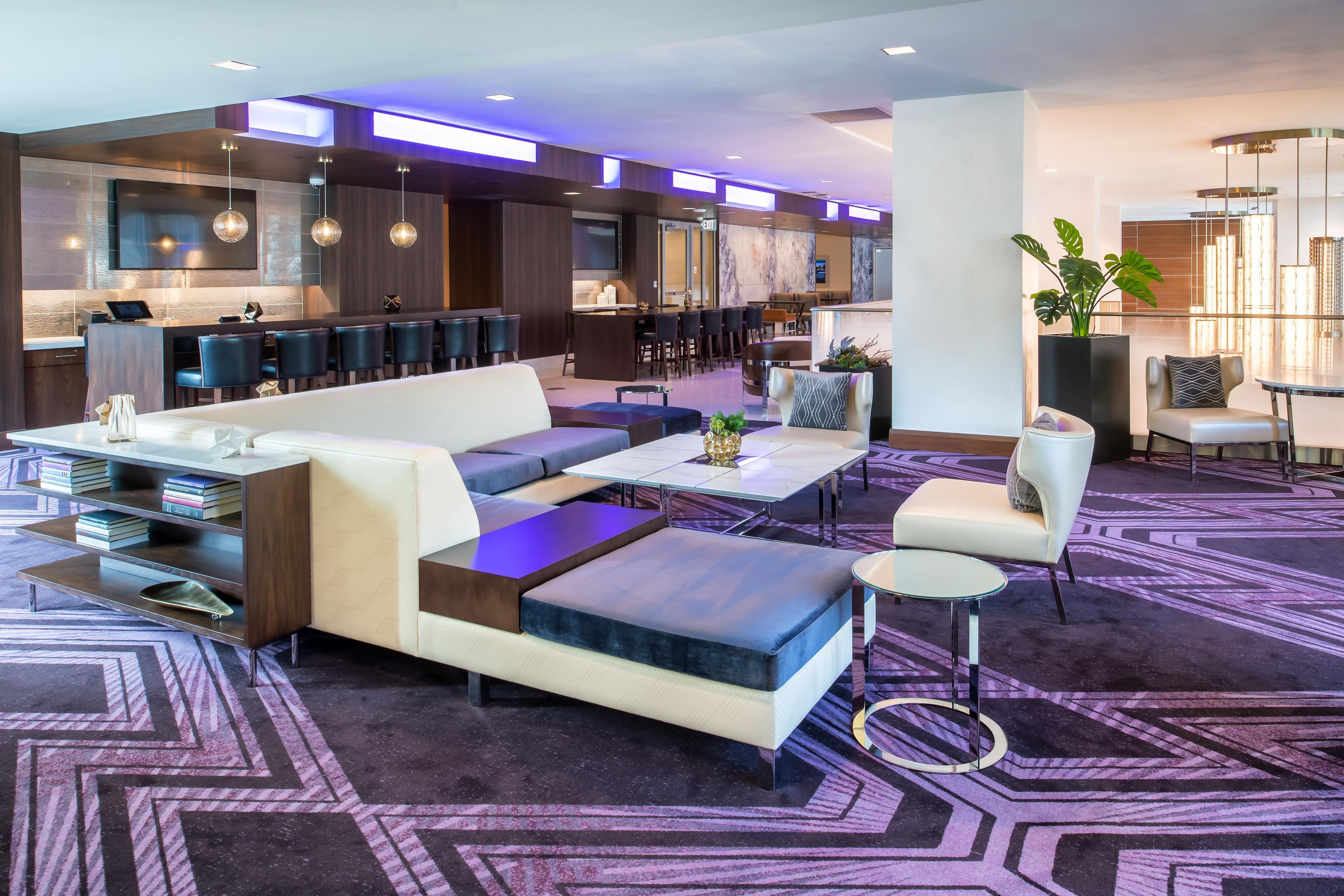 A modern hotel lobby with a sofa, coffee table, and chairs. There is a bar area with stools and a television.