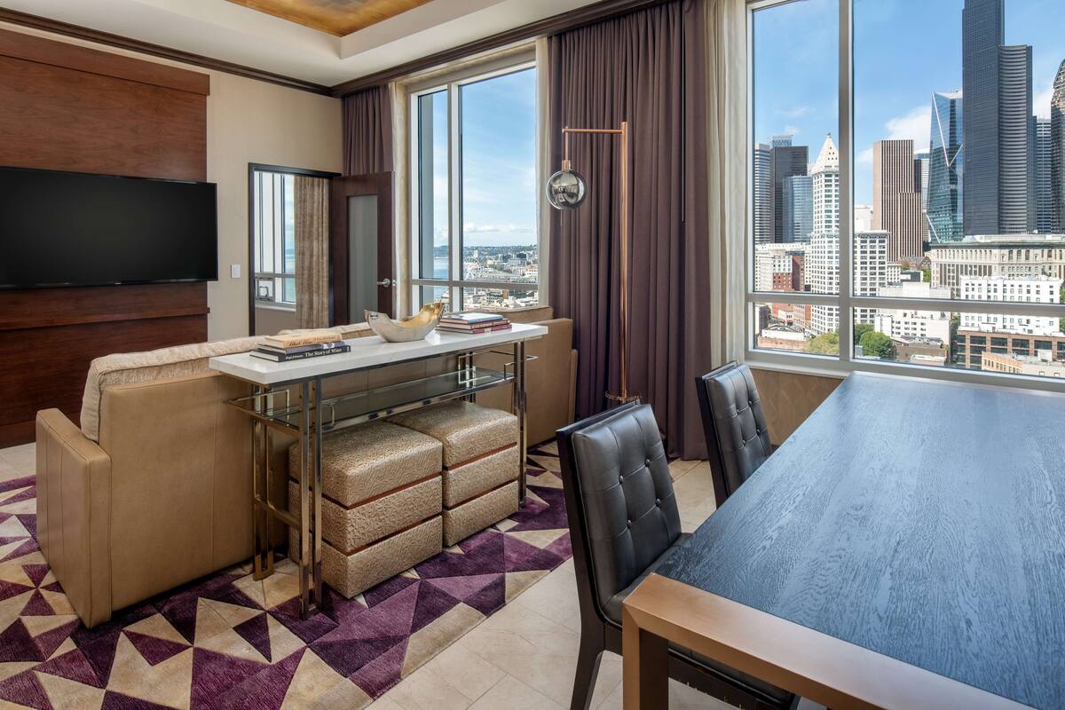 A modern hotel room with a large window offering a city view, a flat-screen TV mounted on the wall, a dining area with a table and chairs, and a seating area with a sofa and ottoman.