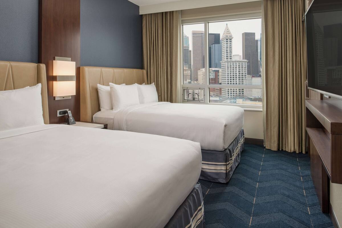 A hotel room with two beds, a large window with a city view, a flat-screen TV, and a wooden nightstand.
