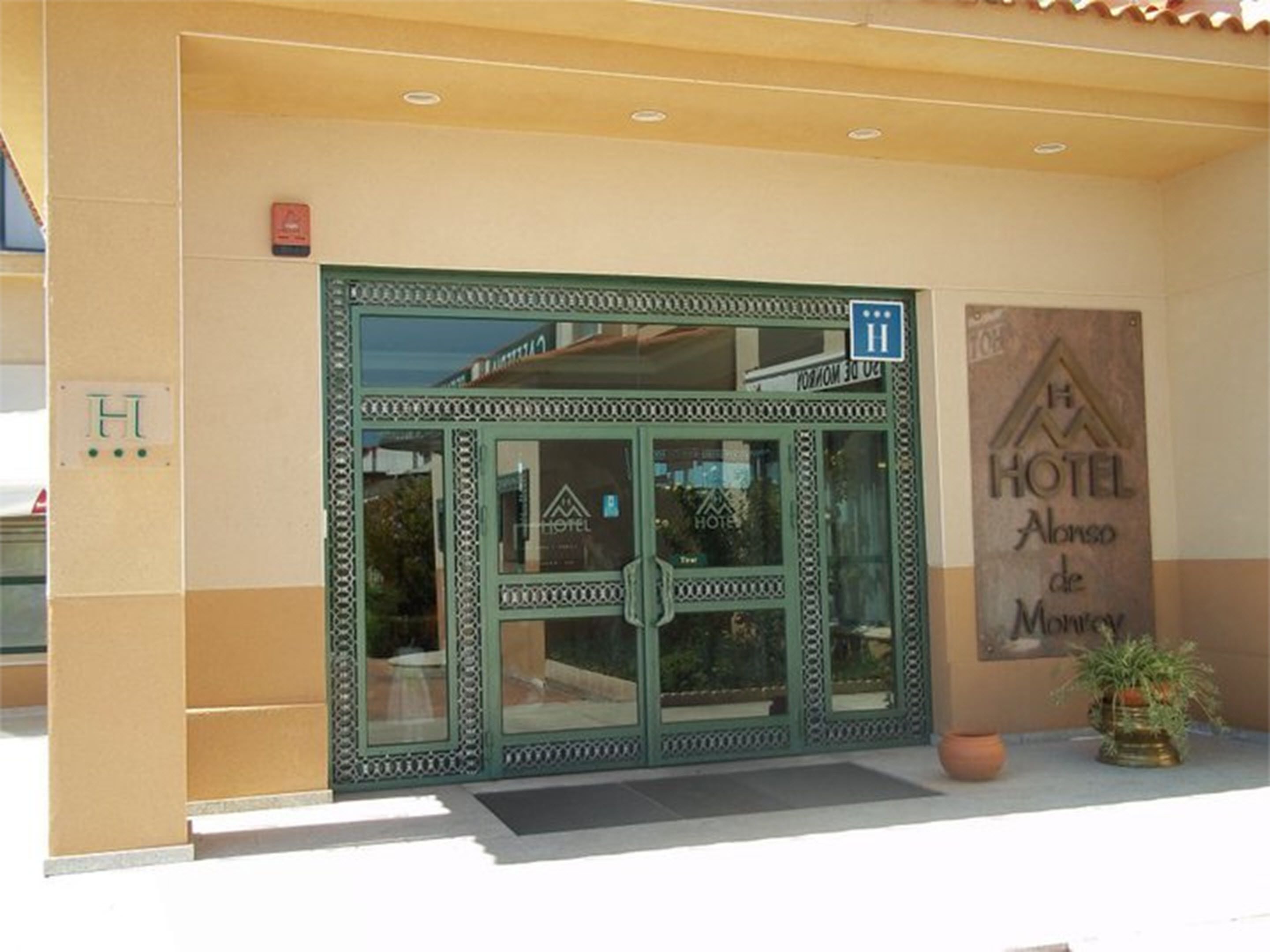 L’entrée d’un hôtel, avec une porte verte, une enseigne indiquant le nom de l’hôtel et un pot de fleurs placé sur la droite.