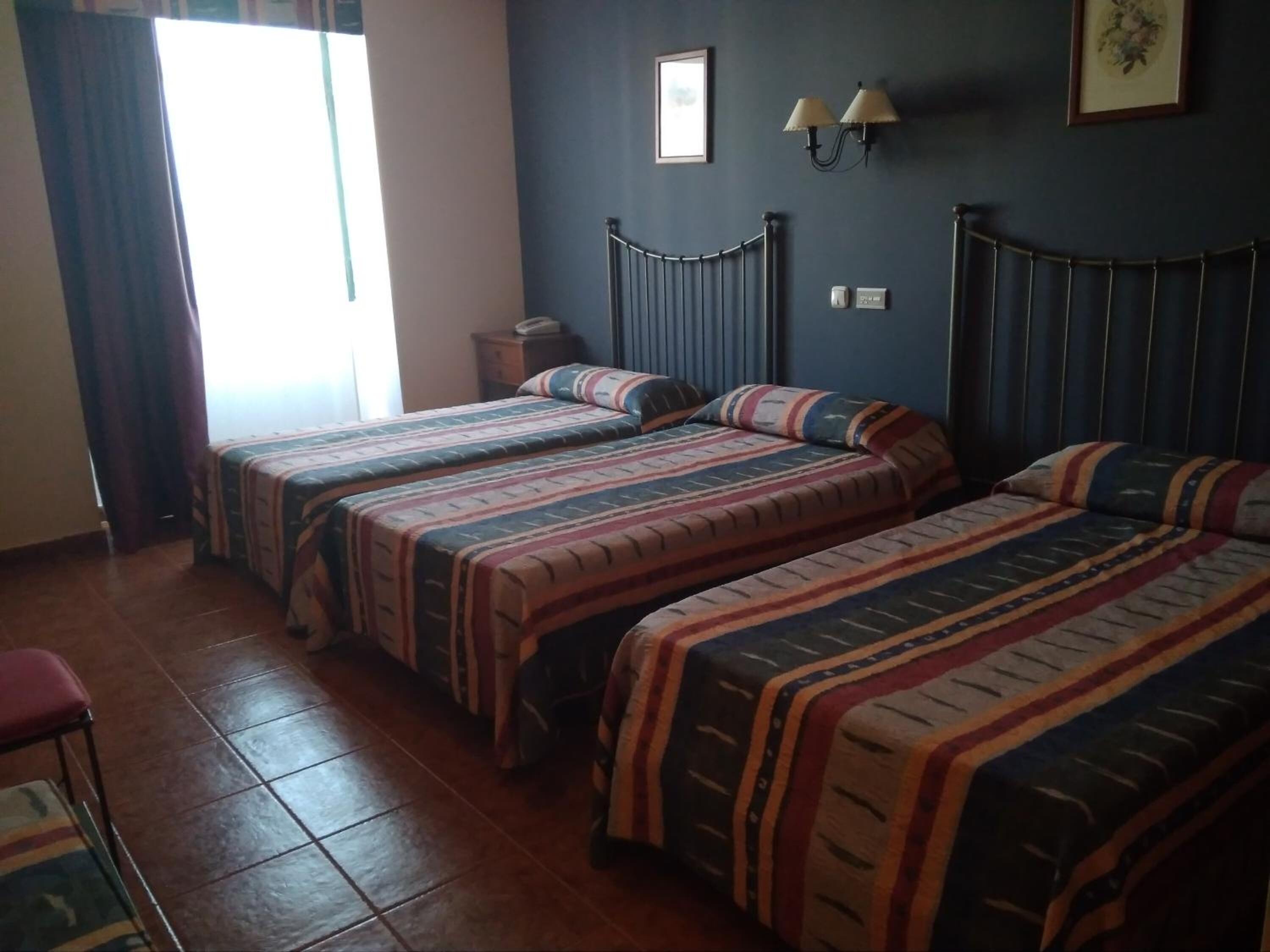 Una habitación con dos camas, una mesita y una silla. Tiene una ventana con cortinas, una lámpara de pared y un cuadro enmarcado.