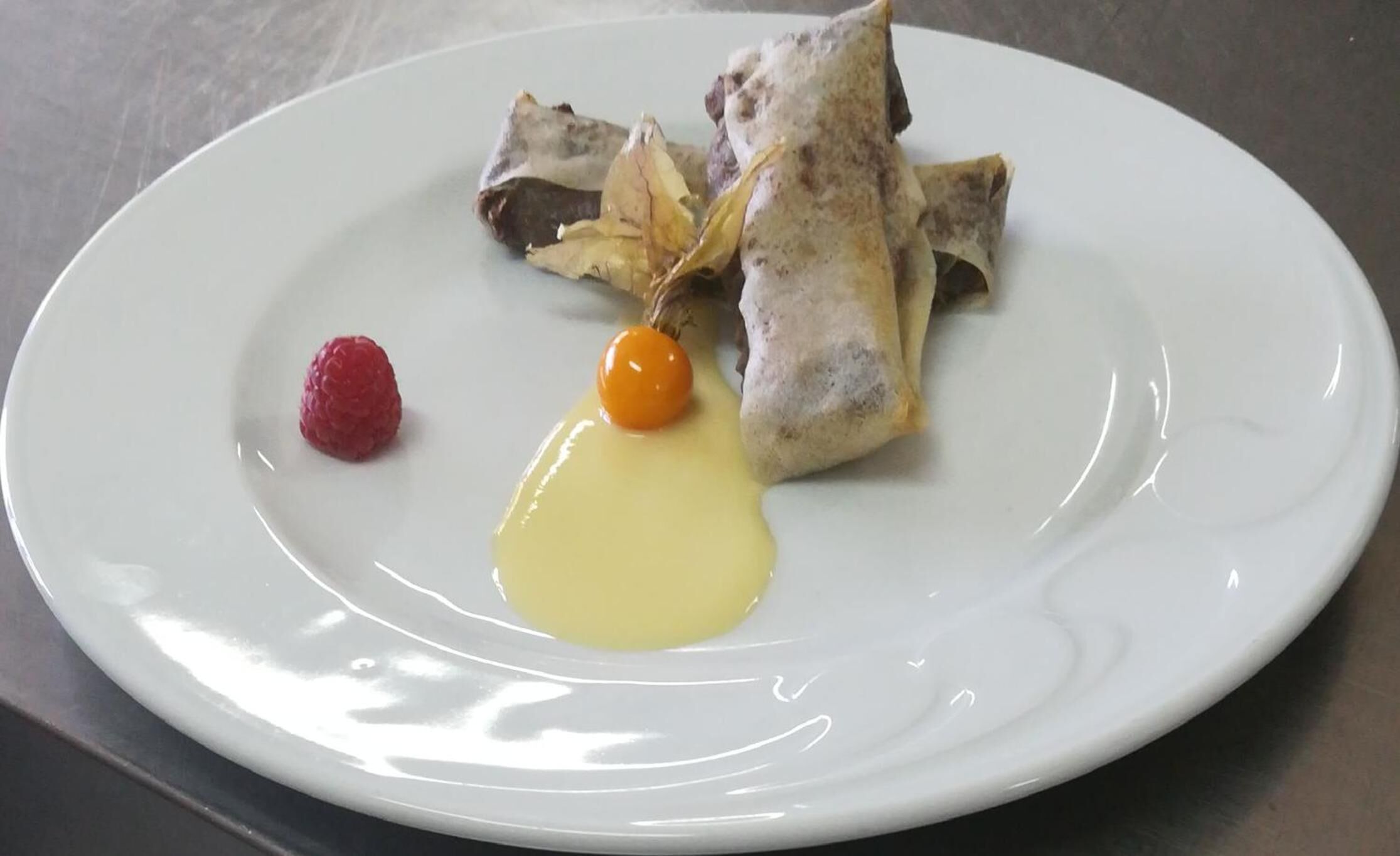 Een wit bord met een dessert bestaande uit een rolletje gebak, een framboos en een lepel gele saus.