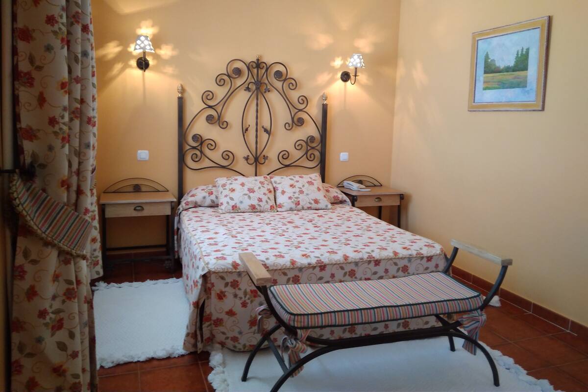Ein Schlafzimmer mit blumigem Bettbezug, einem Metall-Kopfteil, zwei Nachttischlampen, einem Nachttisch, einem Sessel und einem Bild an der Wand.