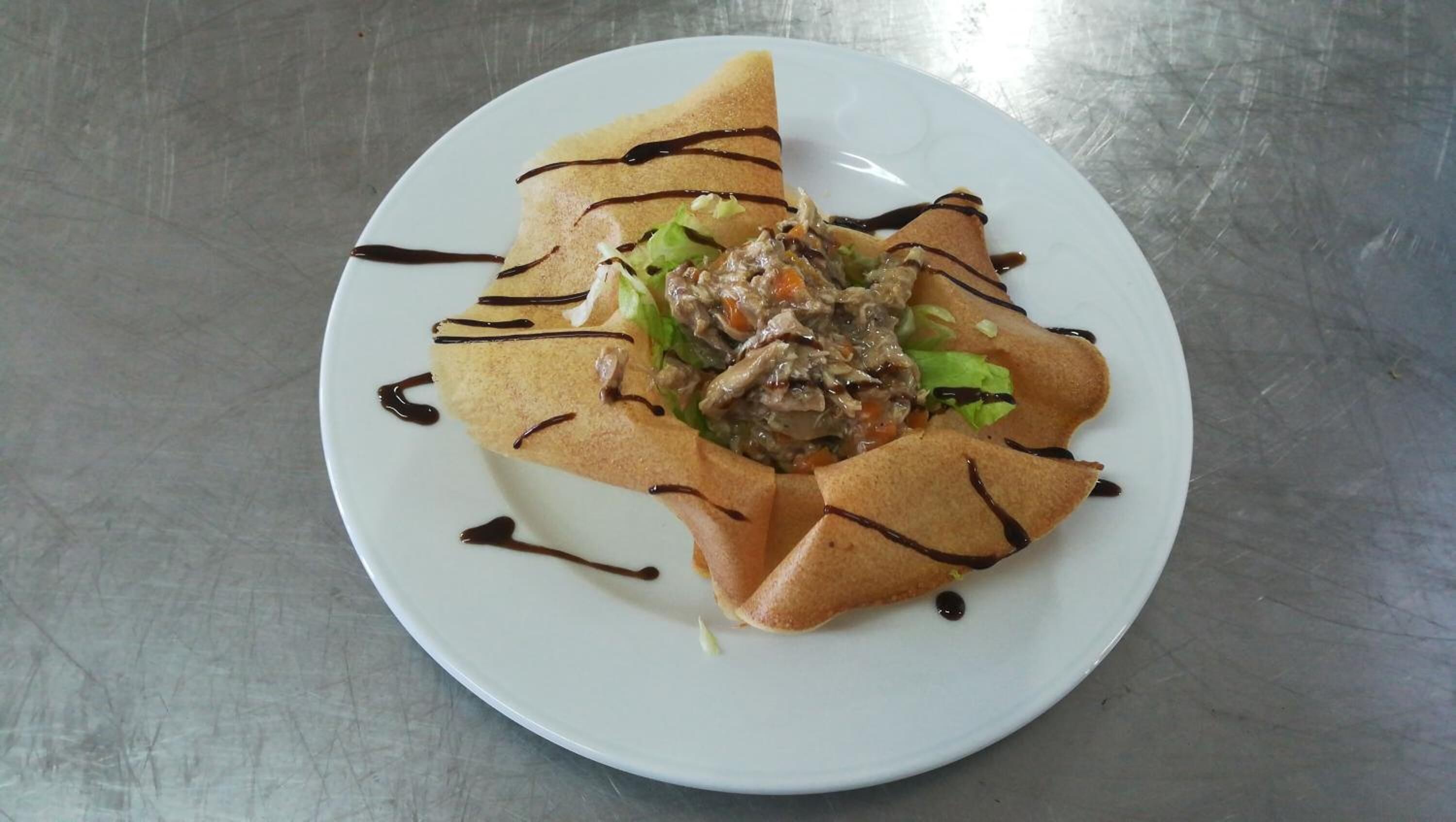A plate with a crepe-like dish topped with shredded meat and vegetables, drizzled with chocolate sauce.