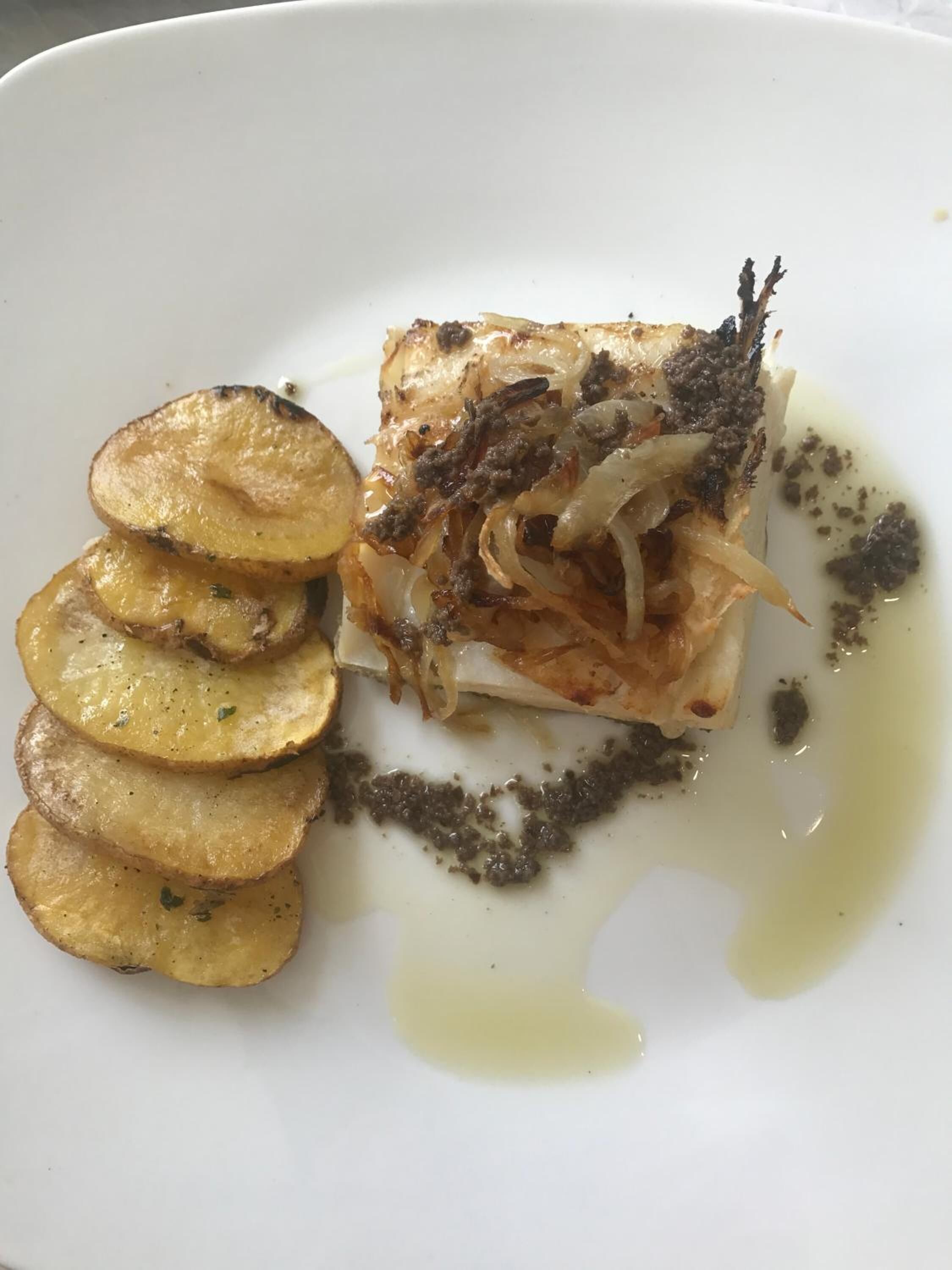 A white plate with a piece of fish topped with onions and a dark sauce, accompanied by roasted potato slices.
