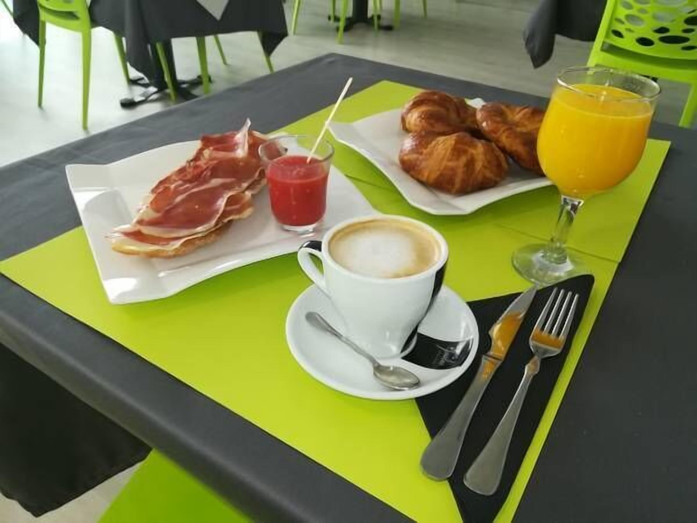 Una mesa de desayuno con cruasanes, un vaso de zumo de naranja, una taza de café y un plato de jamón serrano.