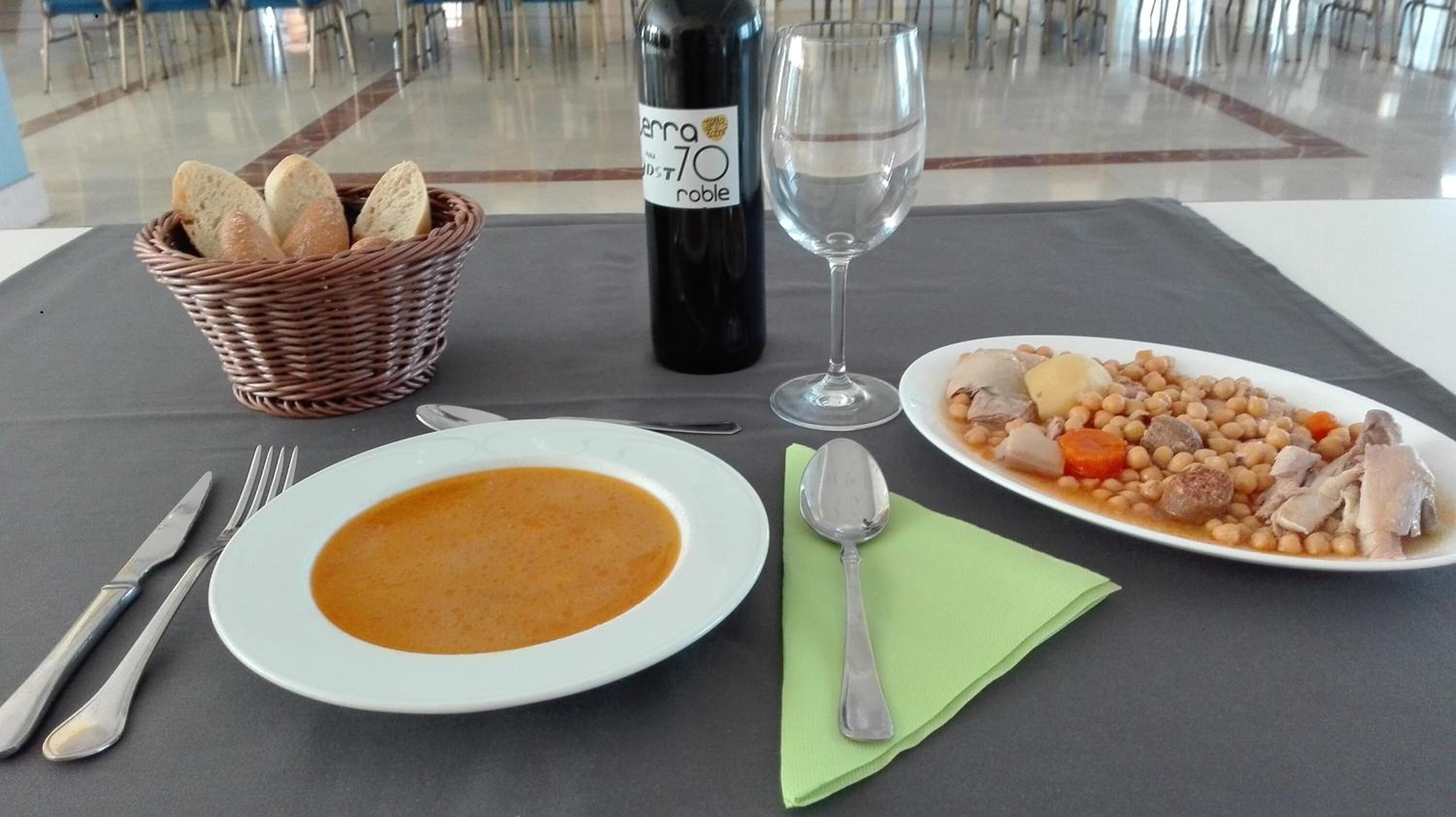 Una mesa servida con un tazón de sopa, un plato de frijoles y carne, una botella de vino y una cesta de pan.