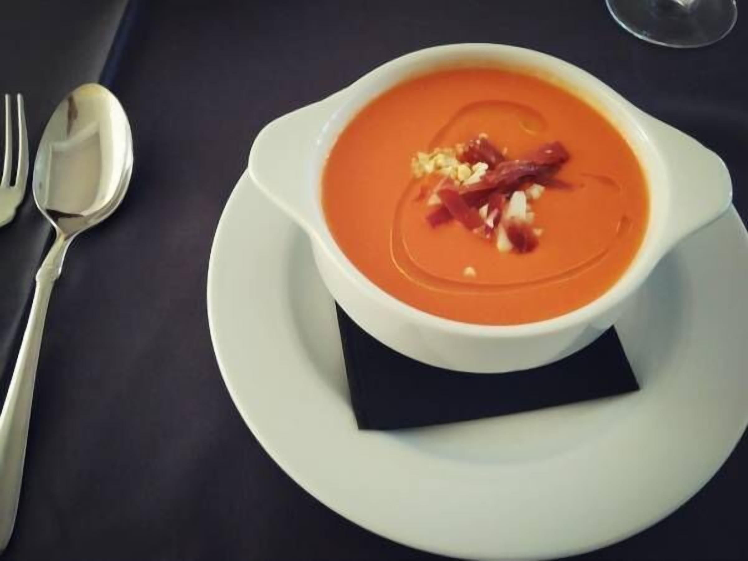 Un tazón de sopa con adorno sobre un plato blanco, junto a un tenedor y una cuchara sobre una mesa oscura.