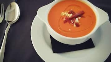 Un tazón de sopa con adorno sobre un plato blanco, junto a un tenedor y una cuchara sobre una mesa oscura.