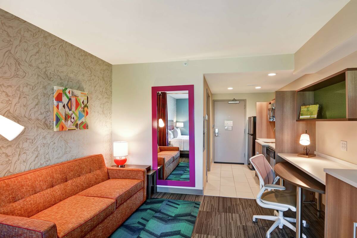 A hotel room with a sofa, a desk, a chair, a bed with a pink frame, and a mirror.