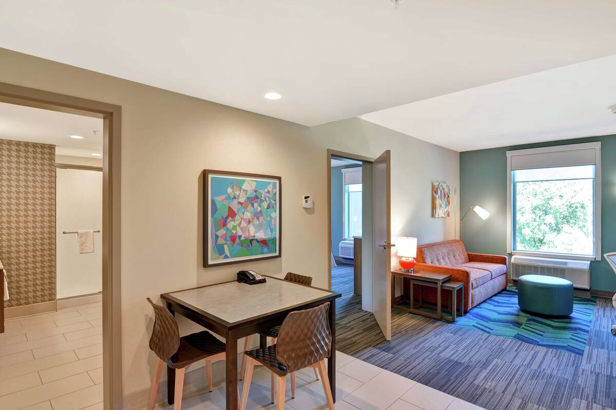 A modern hotel room with a dining area, a sofa, and a large window.