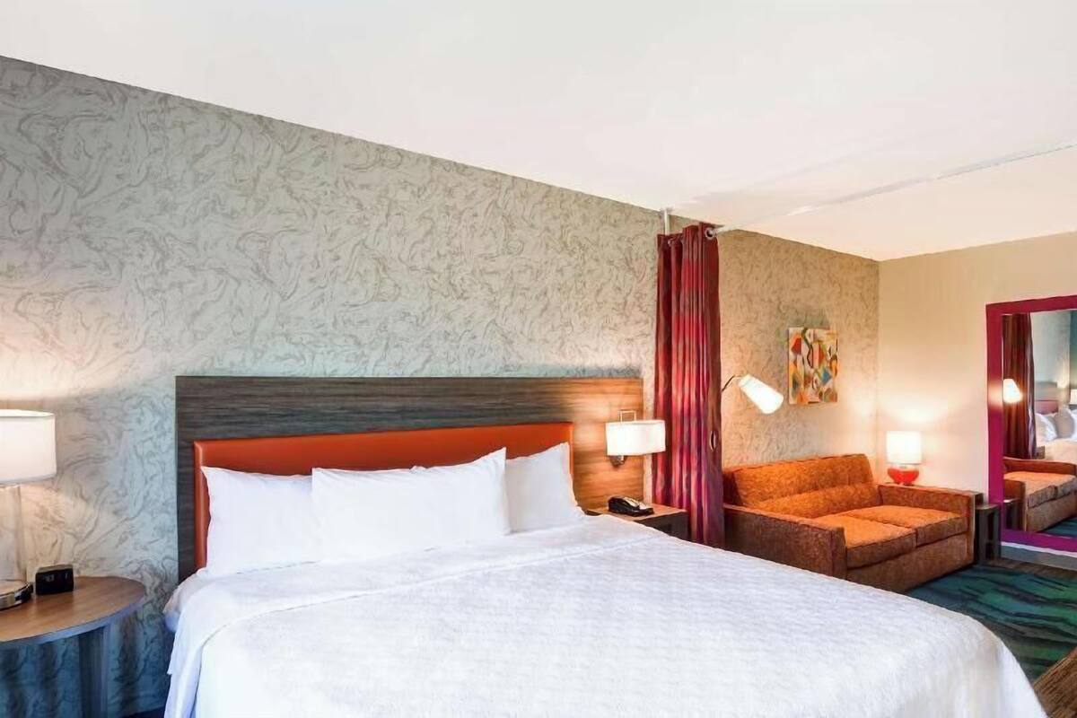 A hotel room with a bed, a nightstand, a chair, a lamp, and a mirror.