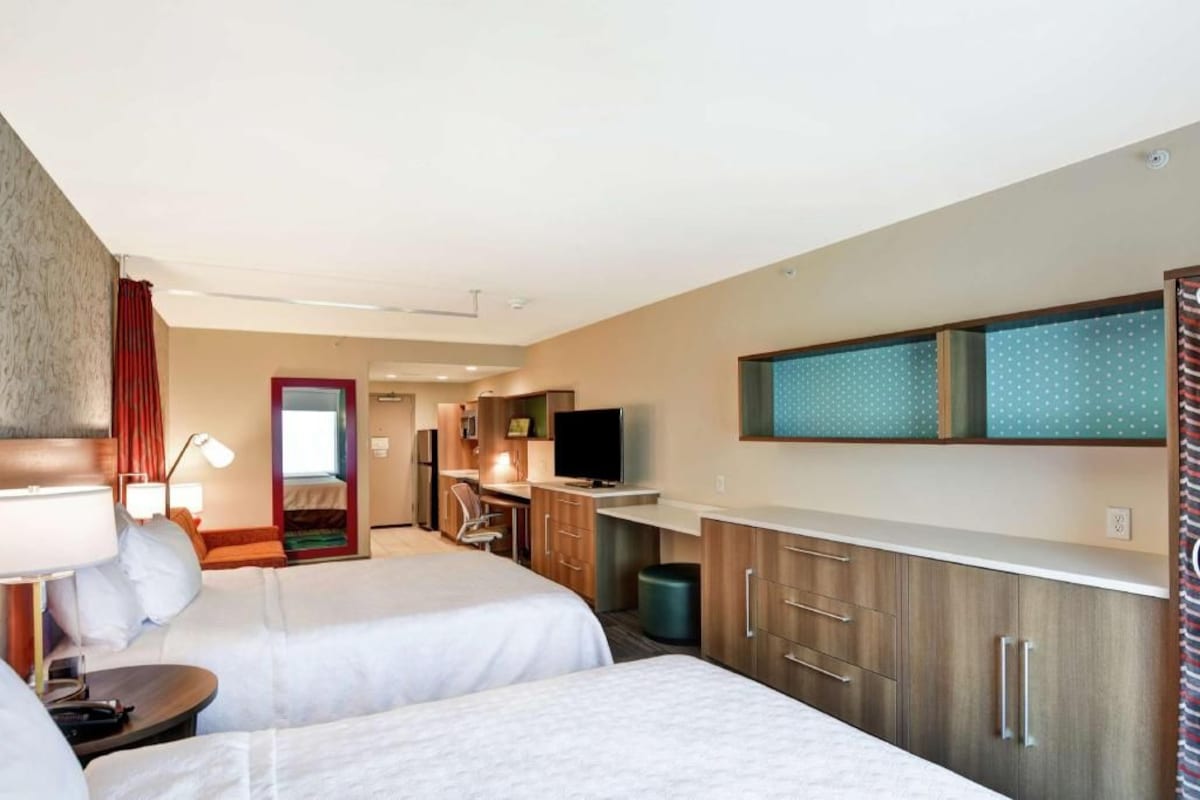 A hotel room with two beds, a desk with a chair, a television, and a kitchenette.
