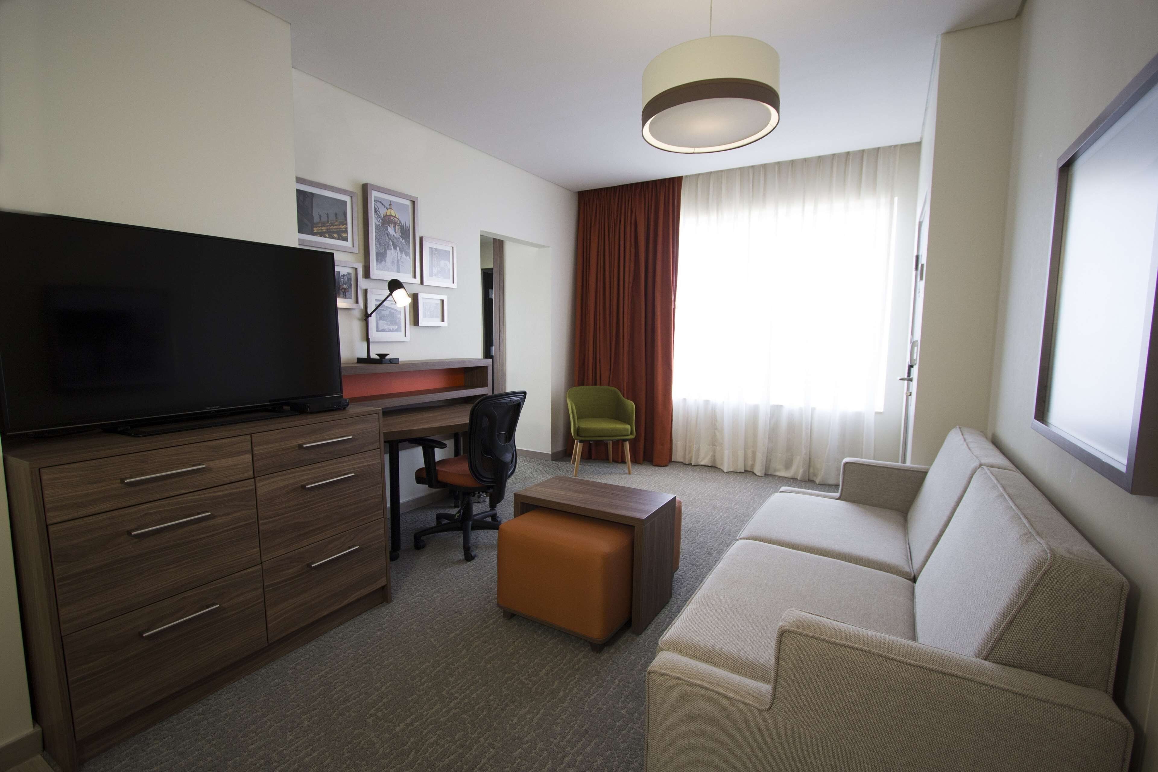 Habitación de hotel con televisor de pantalla plana, un escritorio de madera con cajones, una silla verde, un sofá color beige y una ventana con cortinas.