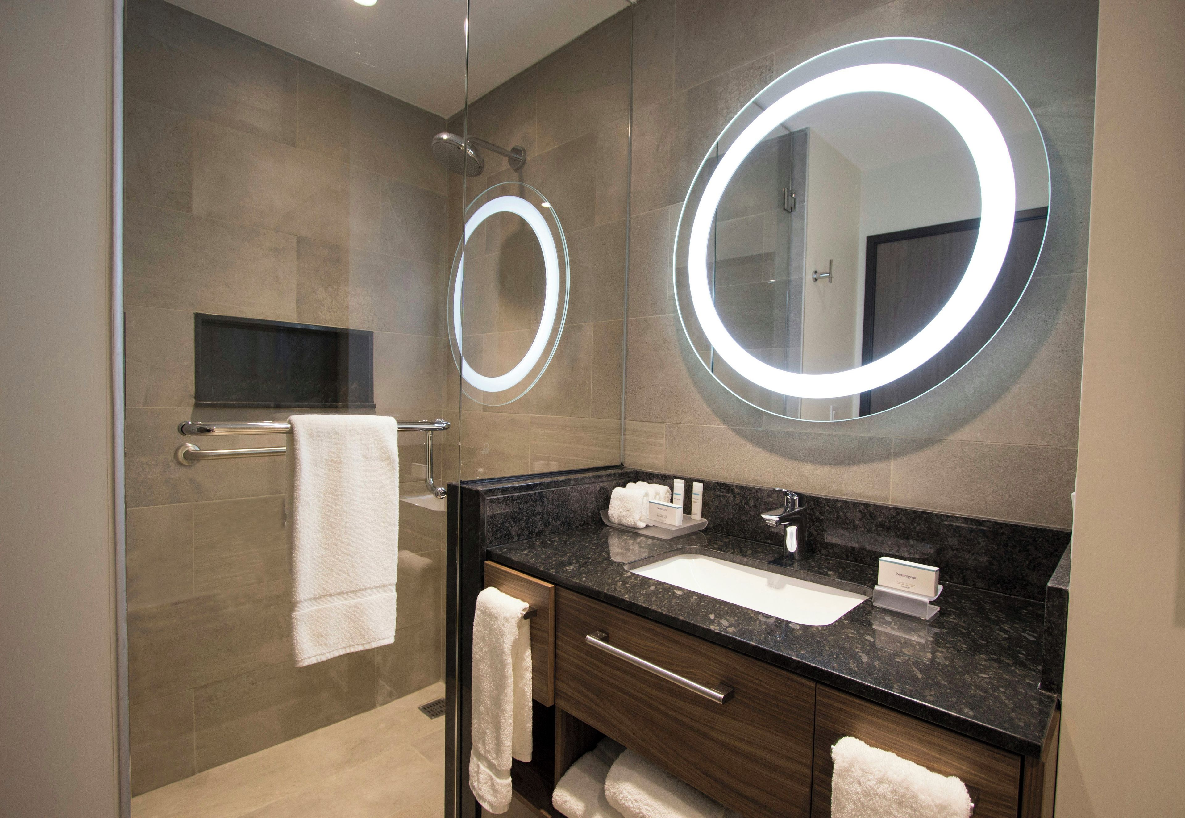 Une salle de bain moderne dotée d’une douche à parois de verre, d’un meuble-lavabo avec un plan de travail en granit foncé et de deux grands miroirs muraux équipés d’un éclairage intégré.