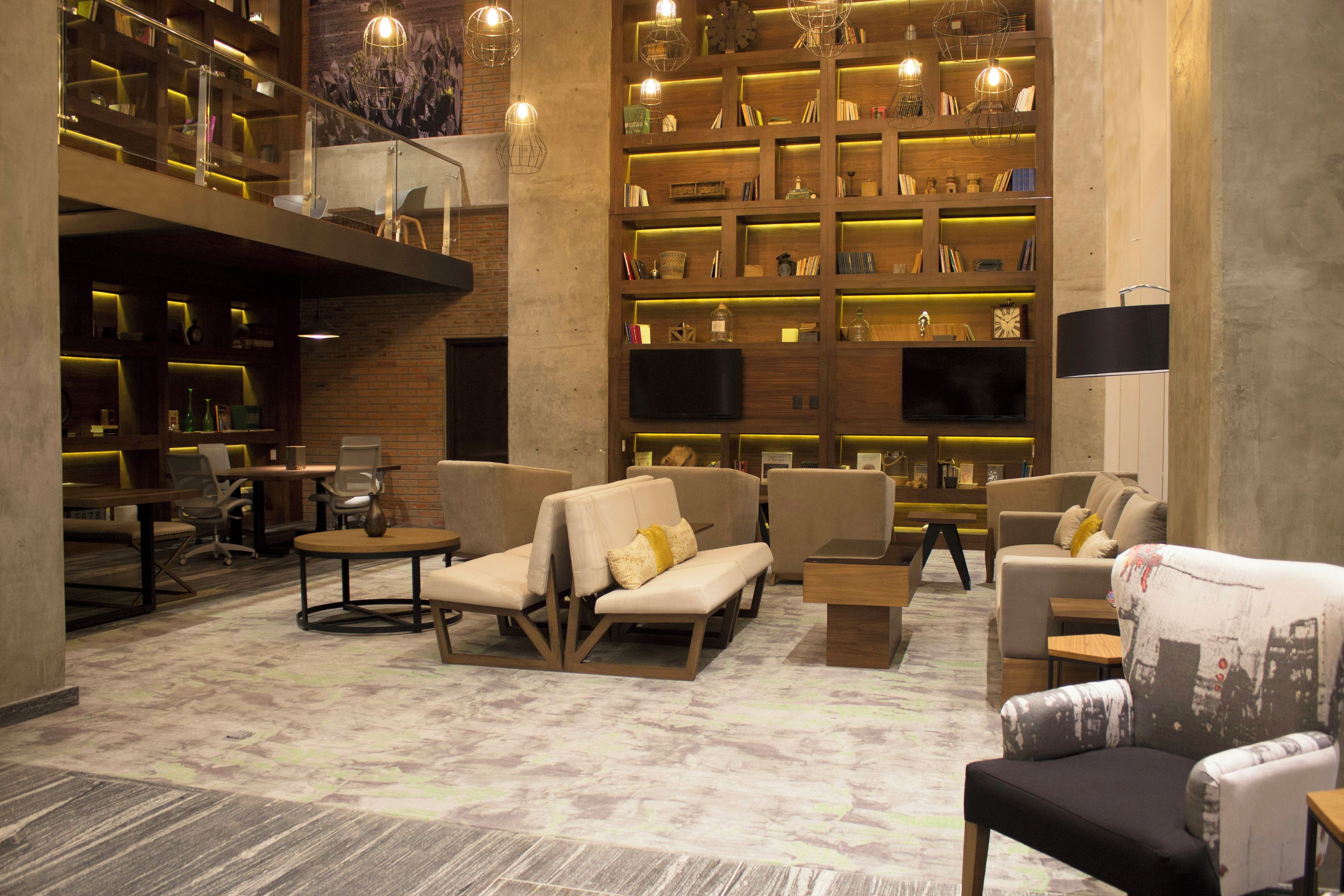 A spacious lobby with a high ceiling, featuring a central seating area with sofas and armchairs, surrounded by bookshelves and a television.