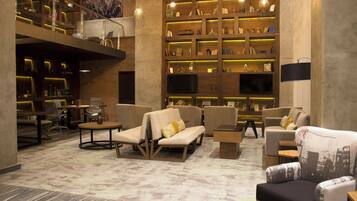 A spacious lobby with a high ceiling, featuring a central seating area with sofas and armchairs, surrounded by bookshelves and a television.