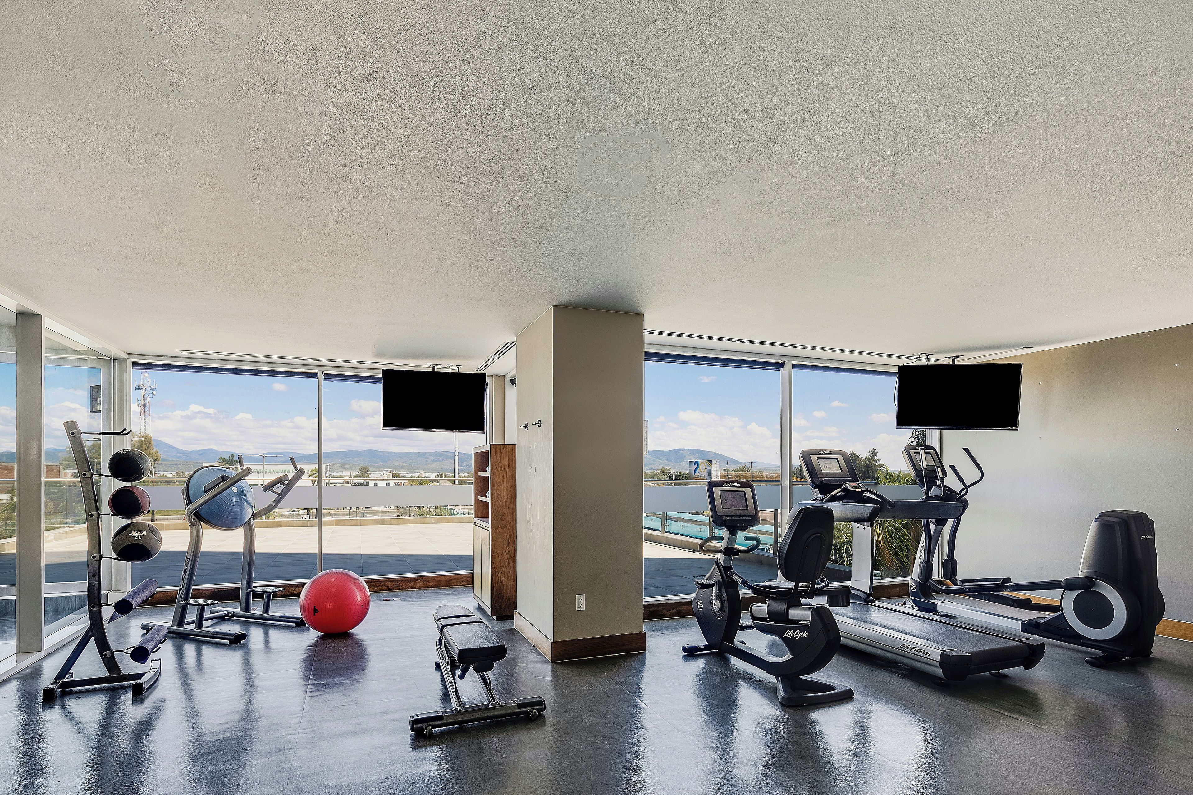 Un gimnasio con equipamiento de ejercicio, amplias ventanas y vistas al exterior.