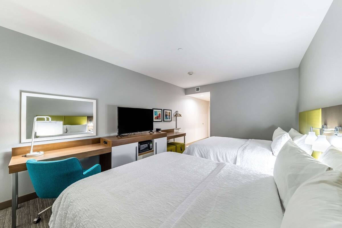 A hotel room with a bed, a desk with a computer, a TV, a mirror, and a chair.