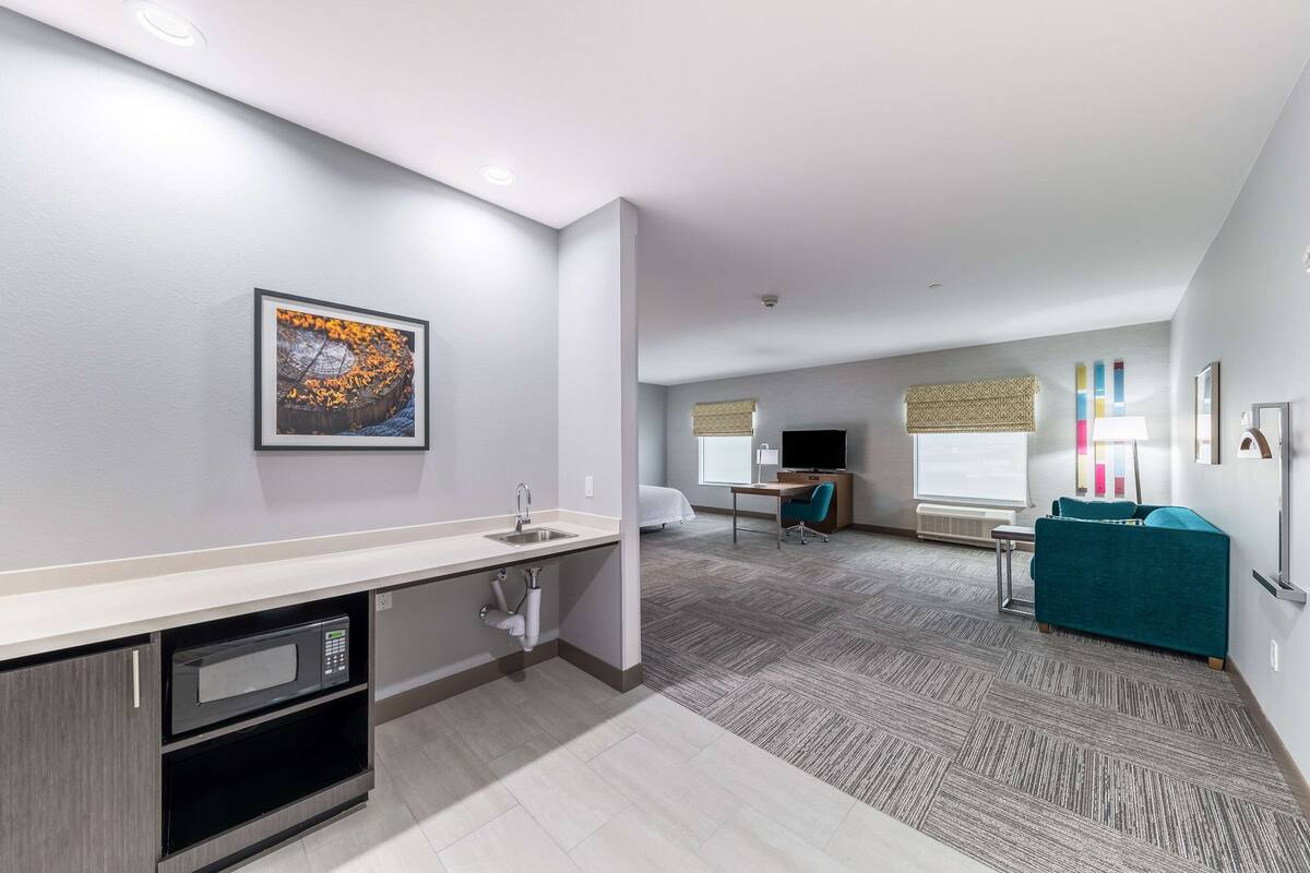 A modern hotel room with a kitchenette, a large window, a desk, and a teal sofa.