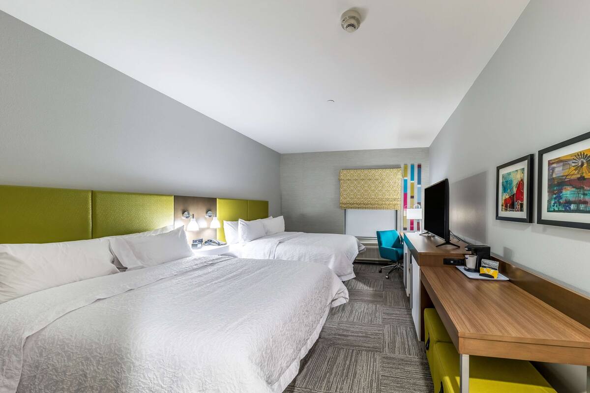A hotel room with two beds, a desk, a chair, a television, and artwork on the wall.
